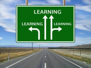 A straight road in the middle of barren land with a road sign above it that has "learning" on it and arrows pointing in different directions