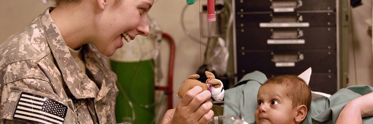 A US military person smiling showing a small stuffed animal to a small child in a patient room with medical equipment in the background