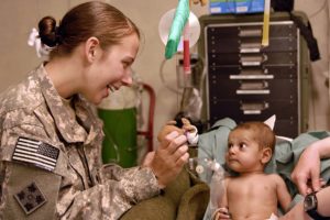 A US military person smiling showing a small stuffed animal to a small child in a patient room with medical equipment in the background