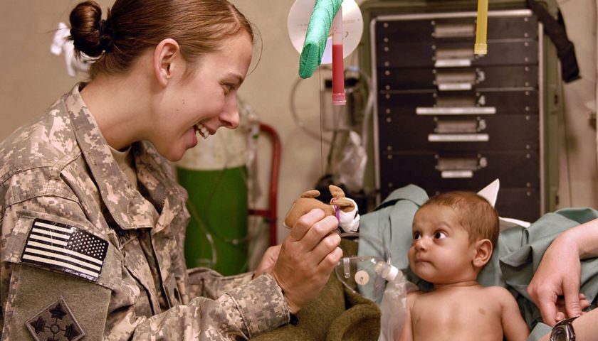 A US military person smiling showing a small stuffed animal to a small child in a patient room with medical equipment in the background