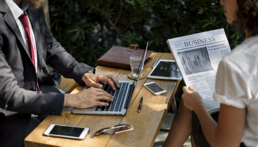 bearded man in black suit, white shirt, red tie working on laptop sitting across from women reading business newspaper at a wood table with cell phones and tablet on table.