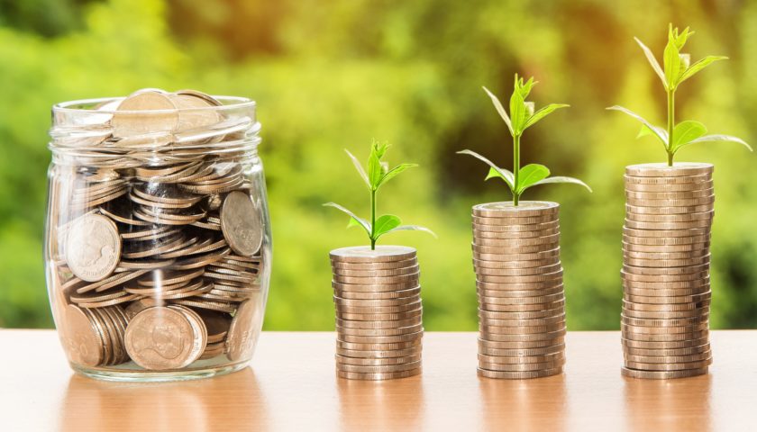 A jar of money with three sets of stacked coins to the right with plants growing out of the top of the conis