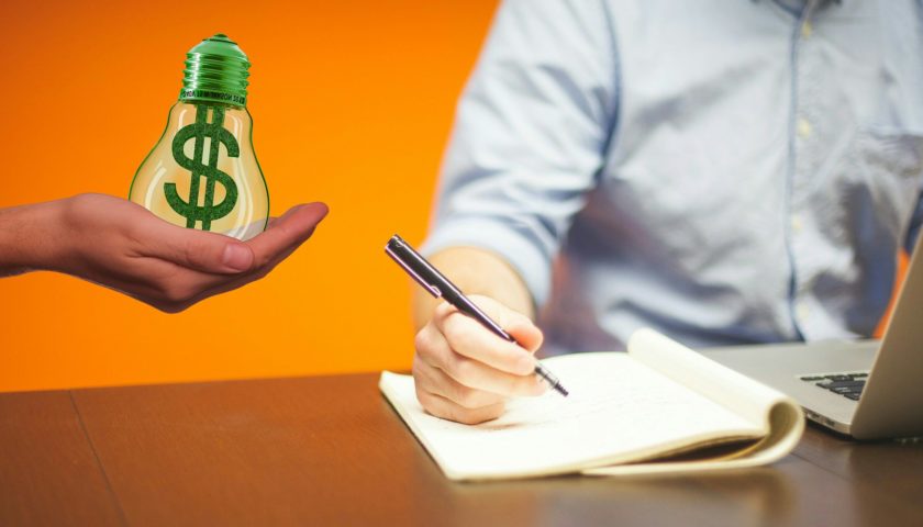 light bulb with green $ with writer in back ground behind a laptop holding a pen on note pad